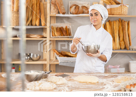 Young female baker whips ingredients in metal bowl Young female baker whips ingredients in metal bowl 128158613