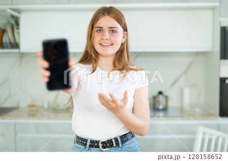 Smiling teenage girl presenting smartphone with blank black screen with copy space in modern cuisine. Free copy space for your design or advertisement 128158652