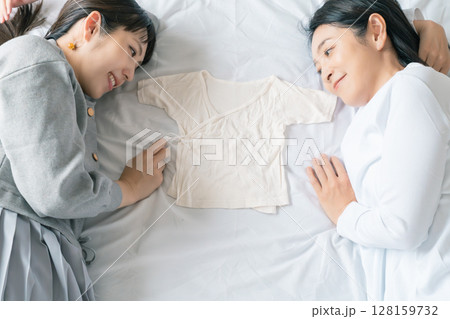 ベッドに寝転び肌着を挟んで赤ちゃんが待ち遠しい母と娘 ベッドに寝転び肌着を挟んで赤ちゃんが待ち遠しい母と娘 128159732