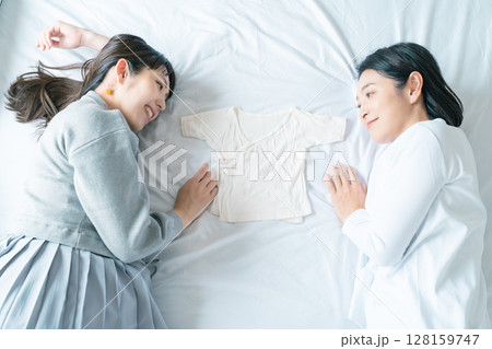 ベッドに寝転び肌着を挟んで赤ちゃんが待ち遠しい母と娘 ベッドに寝転び肌着を挟んで赤ちゃんが待ち遠しい母と娘 128159747