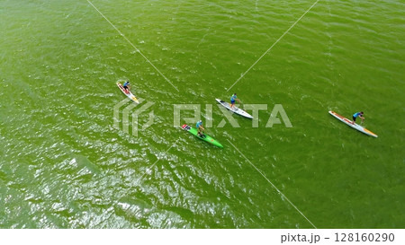 Top down view of four paddleboarders racing on vibrant green water during a stand-up paddleboarding competition with numbered jerseys and paddles. Top down view of four paddleboarders racing on vibrant green water during a stand-up paddleboarding competition with numbered jerseys and paddles. 128160290
