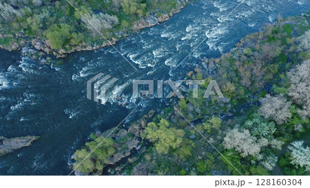 Aerial view of a turbulent flowing river. There are trees and large stone boulders all around. The wind sways the branches of the trees. 128160304