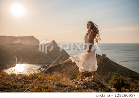 A woman stands on a hill overlooking the ocean. She is wearing a white dress and has long hair. The scene is serene and peaceful, with the sun shining brightly in the background. A woman stands on a hill overlooking the ocean. She is wearing a white dress and has long hair. The scene is serene and peaceful, with the sun shining brightly in the background. 128160957