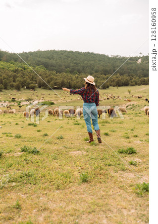A woman in a cowboy hat is standing in a field with a herd of sheep 128160958