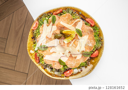 A beautifully plated salmon carpaccio with parmesan shavings, fresh greens, cherry tomatoes, capers, and lemon slices, elegantly arranged on a rustic dish 128161652