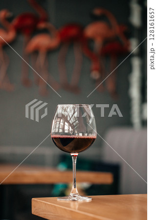 A single glass of red wine stands on a wooden table with a blurred background of pink flamingo decor and cozy indoor seating A single glass of red wine stands on a wooden table with a blurred background of pink flamingo decor and cozy indoor seating 128161657