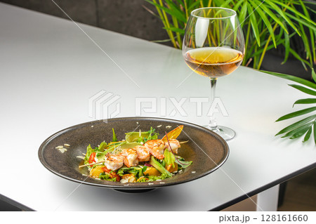 A gourmet shrimp salad with avocado, cherry tomatoes, greens, and microgreens served on a dark plate, paired with a glass of white wine A gourmet shrimp salad with avocado, cherry tomatoes, greens, and microgreens served on a dark plate, paired with a glass of white wine 128161660