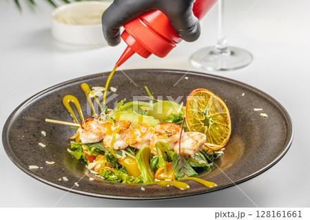 A close-up of a shrimp and vegetable salad being drizzled with bright yellow dressing, garnished with citrus and fresh greens A close-up of a shrimp and vegetable salad being drizzled with bright yellow dressing, garnished with citrus and fresh greens 128161661