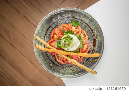 Fresh caprese salad with creamy burrata cheese, ripe tomato slices, green arugula, and crispy breadsticks served on a rustic ceramic plate for a light and elegant appetizer Fresh caprese salad with creamy burrata cheese, ripe tomato slices, green arugula, and crispy breadsticks served on a rustic ceramic plate for a light and elegant appetizer 128161670