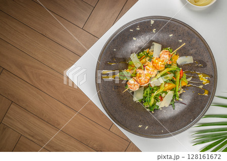 A gourmet shrimp salad beautifully plated with grilled prawns, fresh greens, shaved parmesan, cherry tomatoes, yellow bell peppers, and drizzled with olive oil on a dark ceramic plate 128161672