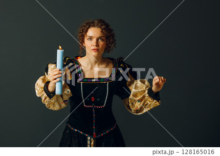 Portrait of a young adult woman dressed in a medieval dress holds a candle on dark background 128165016