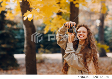 Smiling young woman photographer with the photo camera shooting the forest trees with the yellow leaves at sunset 128165020