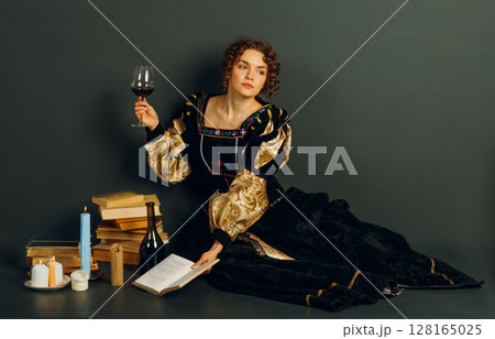 Young adult woman dressed in a medieval dress sitting near ancient books and candles with bottle of red wine and holding a glass of wine Young adult woman dressed in a medieval dress sitting near ancient books and candles with bottle of red wine and holding a glass of wine 128165025