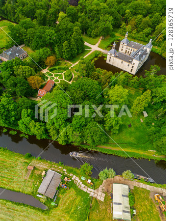 Aerial view of a historic castle surrounded by a forest, with vast green fields, winding waterways, and a river stretching across the flat Dutch countryside under a partly cloudy sky. Aerial view of a historic castle surrounded by a forest, with vast green fields, winding waterways, and a river stretching across the flat Dutch countryside under a partly cloudy sky. 128165719