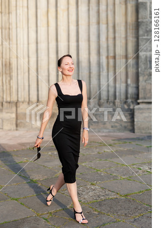 Portrait young adult elegant woman black dress and sunglasses smiles confidently in golden sunlight warm sunset backlit light against historic cathedral building. Female person walking city street Portrait young adult elegant woman black dress and sunglasses smiles confidently in golden sunlight warm sunset backlit light against historic cathedral building. Female person walking city street 128166161