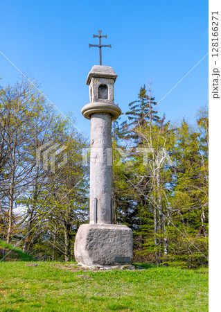 A tall stone column with a cross at the top rises from a lush green field surrounded by trees. The sky is clear and bright, emphasizing the peaceful yet haunting presence of the structure. A tall stone column with a cross at the top rises from a lush green field surrounded by trees. The sky is clear and bright, emphasizing the peaceful yet haunting presence of the structure. 128166271