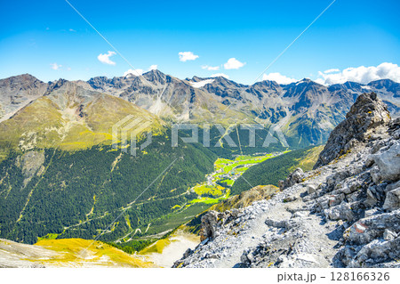 A panoramic view of the Sulden Valley in Italy, showcasing lush greenery amidst rugged mountains under a clear blue sky during summer. 128166326