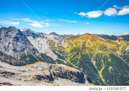 The winding roads of Stelvio Pass cut through the Italian Alps, showcasing a blend of rugged peaks and summer greenery against a clear blue sky. 128166327