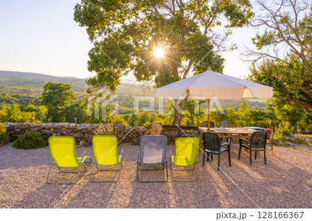 Enjoy a peaceful summer sunset as golden rays filter through trees, illuminating a cozy outdoor space with chairs and a table, perfect for gathering with friends or family. 128166367