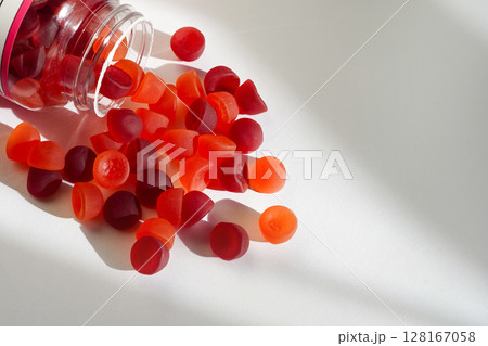 A transparent bottle lies on its side as colorful red and orange gummy vitamins spill onto a clean white surface A transparent bottle lies on its side as colorful red and orange gummy vitamins spill onto a clean white surface 128167058