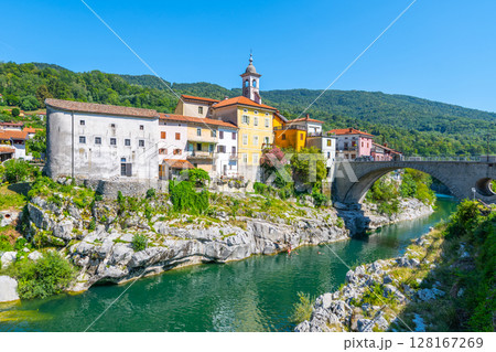 Kanal bridge gracefully stretches over the clear waters of the Soca River, surrounded by charming houses nestled against a backdrop of lush green hills on a sunny day. 128167269