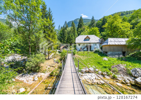 Hikers traverse a wooden bridge spanning Vrsnica creek in the Soca Valley. Lush greenery surrounds them, highlighting the beauty of the Julian Alps on a sunny day. 128167274