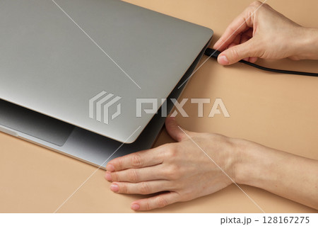 female hand connecting charger cable to laptop at beige table, closeup female hand connecting charger cable to laptop at beige table, closeup 128167275