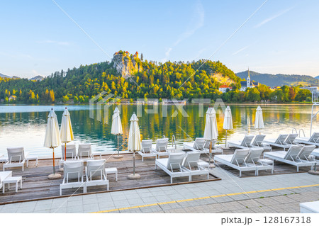 Bled Lake showcases its tranquil waters reflecting the surrounding nature, while Bled Castle stands majestically atop the hill. Lounge chairs await visitors by the lakeside. 128167318