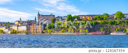 Stockholm showcases its stunning waterfront, featuring historic buildings and lush greenery under a clear blue sky. Boats are docked along the serene water, reflecting the vibrant surroundings. Stockholm showcases its stunning waterfront, featuring historic buildings and lush greenery under a clear blue sky. Boats are docked along the serene water, reflecting the vibrant surroundings. 128167354