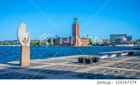 A vibrant day in Stockholm showcases a striking sculpture near the waterfront. The city hall stands prominently, framed by lush trees against a clear blue sky. 128167374