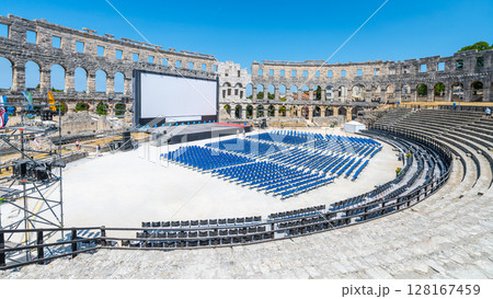The Roman amphitheatre in Pula, Croatia, hosts an outdoor event setup with rows of seating and a large screen. Sunlight bathes the ancient stone structure, highlighting its history and beauty. 128167459