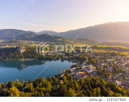 Bled Lake sparkles under the golden sunlight, framed by green forests and mountains. The charming town of Bled is visible, with its historic castle perched on a cliff. Bled Lake sparkles under the golden sunlight, framed by green forests and mountains. The charming town of Bled is visible, with its historic castle perched on a cliff. 128167488