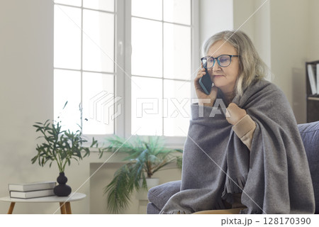 Serious senior woman covered in cozy blanket sitting on home sofa and calling on mobile phone Serious senior woman covered in cozy blanket sitting on home sofa and calling on mobile phone 128170390