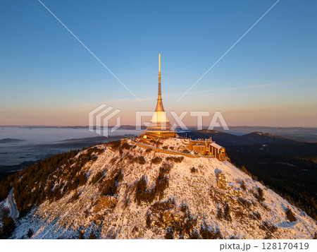 As the sun rises over Jested Mountain, the snow-covered summit glows in warm light, illuminating the surrounding forest and hills. A tranquil winter scene unfolds in the morning mist. 128170419