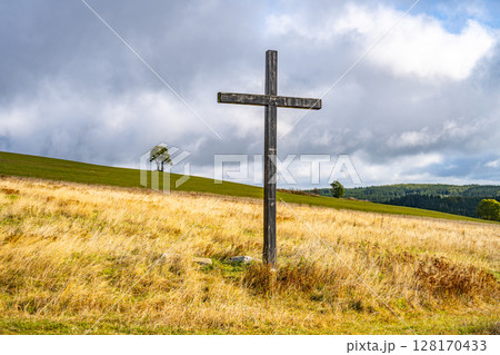 A wooden cross rises prominently in a mountain meadow near Haj, Czechia. Rolling hills and a solitary tree frame this serene landscape under a partly cloudy sky. 128170433