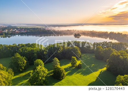 An enchanting aerial perspective of Trebon reveals the town shrouded in morning haze, with serene waters reflecting the soft light. Lush greenery borders the tranquil lake, offering a picturesque 128170443