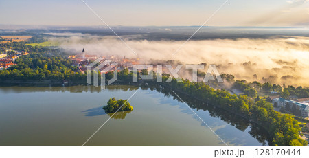 A serene aerial perspective reveals Trebon shrouded in morning mist. The tranquil water reflects the soft haze, creating an ethereal atmosphere over the picturesque Czechia town, awakening with the 128170444