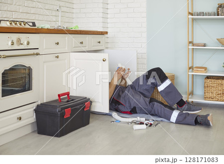 Male plumber in work uniform lying on floor, fixing kitchen sink pipes, providing plumbing service. 128171083