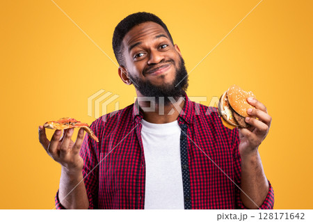 Smiling African Man Holding Burger And Slice Of Pizza Posing Standing In Studio Over Yellow Background. Junk Food Eater's Portrait. Unhealthy Fastfood And Male Nutrition, Cheat Meal And Overeating Smiling African Man Holding Burger And Slice Of Pizza Posing Standing In Studio Over Yellow Background. Junk Food Eater's Portrait. Unhealthy Fastfood And Male Nutrition, Cheat Meal And Overeating 128171642