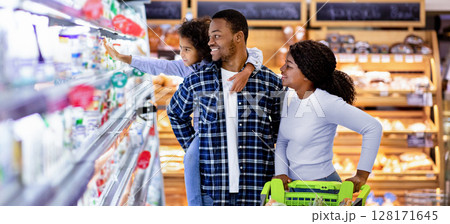 Portrait of happy black family with trolley shopping together at grocery store. Millennial African American parents with lovely daughter picking food, choosing milk products at big mall 128171645