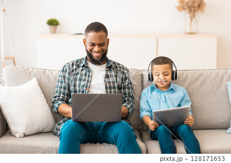 Black Father And Little Boy Using Computers Together Browsing Internet On Laptop And Digital Tablet Wearing Wireless Headphones Sitting On Sofa At Home. Family Gadgets Concept Black Father And Little Boy Using Computers Together Browsing Internet On Laptop And Digital Tablet Wearing Wireless Headphones Sitting On Sofa At Home. Family Gadgets Concept 128171653