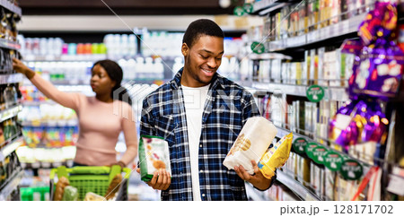 Black man and his girlfriend shopping for groceries, making selection of healthy food at mall, copy space. Positive African American couple buying products at modern supermarket 128171702