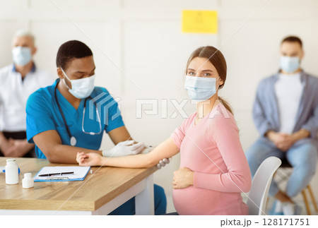 Pregnant woman in face mask getting vaccinated against Covid-19 at clinic. African American doctor giving coronavirus vaccine injection to expectant lady. Healthcare and global immunization 128171751