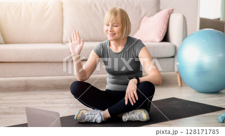 Positive mature woman in sportswear sitting on yoga mat in front of laptop, greeting her personal trainer before training. Senior lady communicating with fitness instructor, ready for home workout Positive mature woman in sportswear sitting on yoga mat in front of laptop, greeting her personal trainer before training. Senior lady communicating with fitness instructor, ready for home workout 128171785