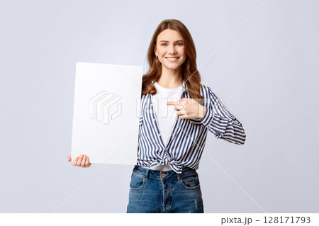 Smiling Millennial Lady Holding Blank White Placard And Pointing At It, Young Caucasian Woman Holding Poster With Copy Space For Your Design Or Advertisement, Standing On Light Background 128171793