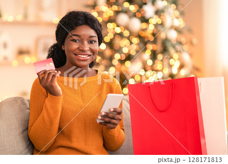 Online shopping and home delivery during winter holidays. Young happy african american woman holds phone and credit card, sits on sofa with paper bags with purchases in living room with Christmas tree Online shopping and home delivery during winter holidays. Young happy african american woman holds phone and credit card, sits on sofa with paper bags with purchases in living room with Christmas tree 128171813