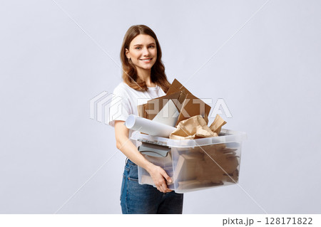 A young woman smiling while holding a transparent container filled with recyclable materials like cardboard and paper. She stands against a light background, promoting eco-friendly practices. A young woman smiling while holding a transparent container filled with recyclable materials like cardboard and paper. She stands against a light background, promoting eco-friendly practices. 128171822