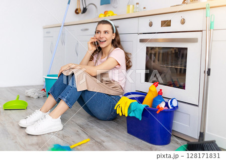 Cheerful young woman speaking on cellphone during break from housework, enjoying phone conversation with friend at kitchen. Happy housewife resting from domestic duties on floor 128171831