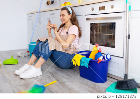 Happy young housewife taking break from home cleaning, browsing internet on smartphone while sitting on kitchen floor. Positive millennial lady checking social media during cleanup Happy young housewife taking break from home cleaning, browsing internet on smartphone while sitting on kitchen floor. Positive millennial lady checking social media during cleanup 128171878