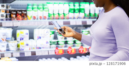 Happy black lady checking her smartphone shopping app while purchasing groceries at dairy section of big mall, copy space. Young African American woman buying foodstuff at supermarket 128171906